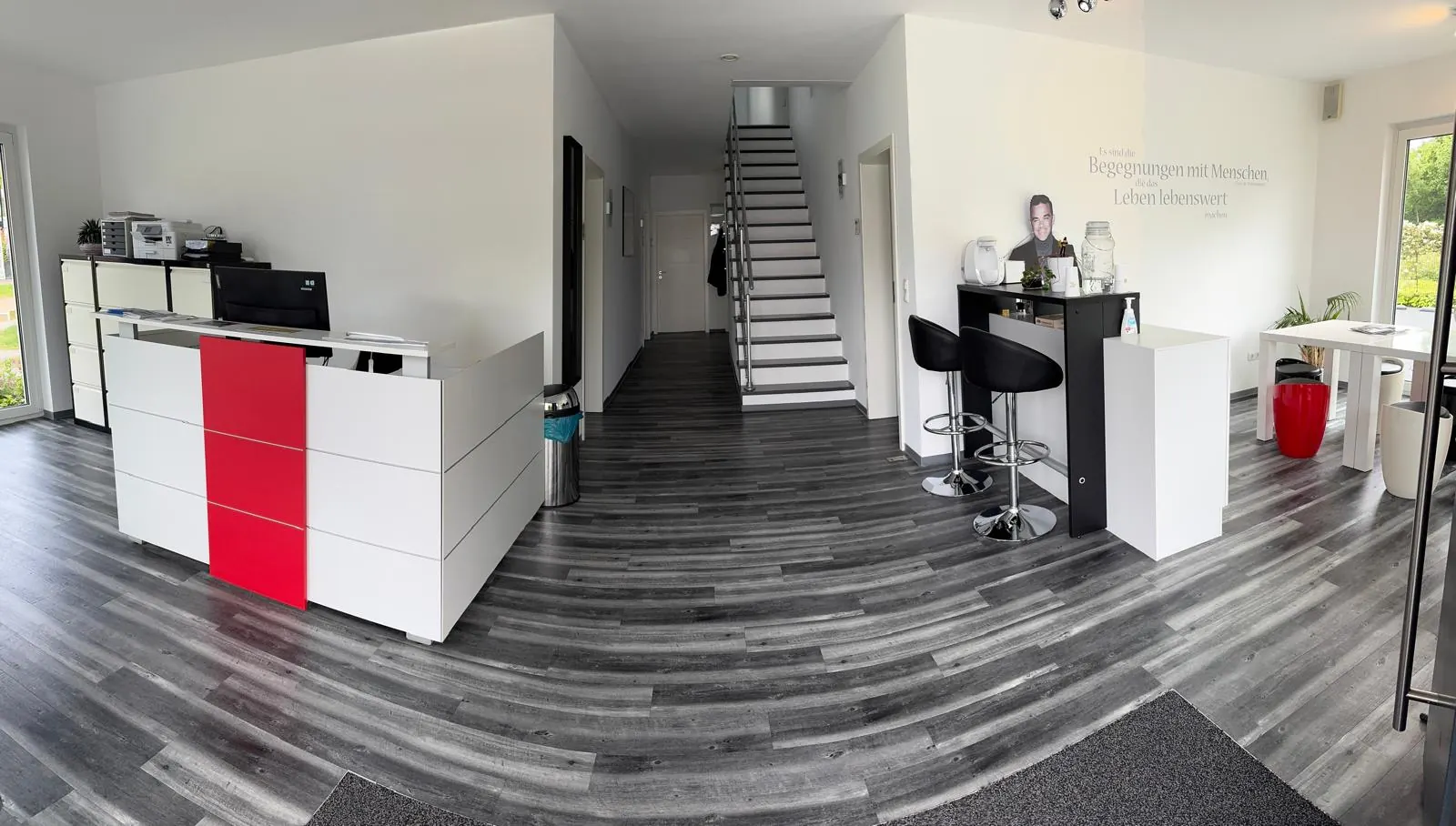 Empfangsbereich des Büros mit weißem Tresen, rotem Farbakzent, Barhockern und Treppe